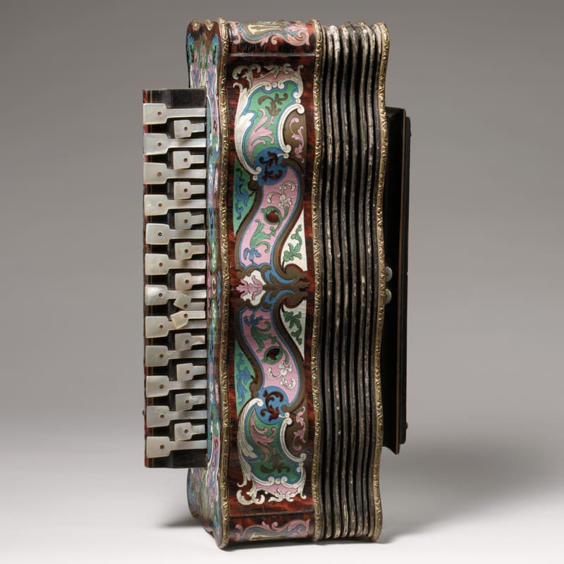 Diatonic button accordion with a tortoiseshell-and-enamel case decorated in pink, green and blue scrollwork, pearl keys along the left side and dark leather bellows, photographed from the side against a grey studio backdrop.