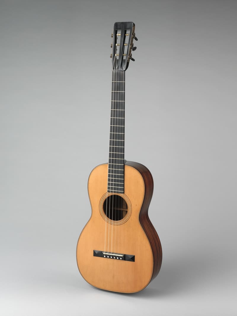 Small parlor-style acoustic guitar with a natural spruce top, rosewood sides, black ebony fingerboard and slotted headstock with side-mounted tuners, standing at a three-quarter angle against a grey backdrop.