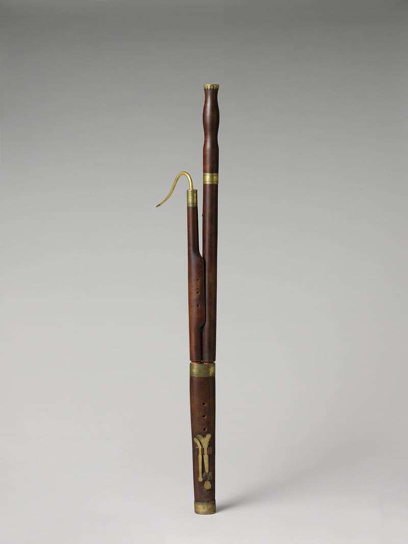 A wooden bassoon in four sections with brass bands, a curved brass bocal and one key at the bell, shown upright against a soft grey studio background.