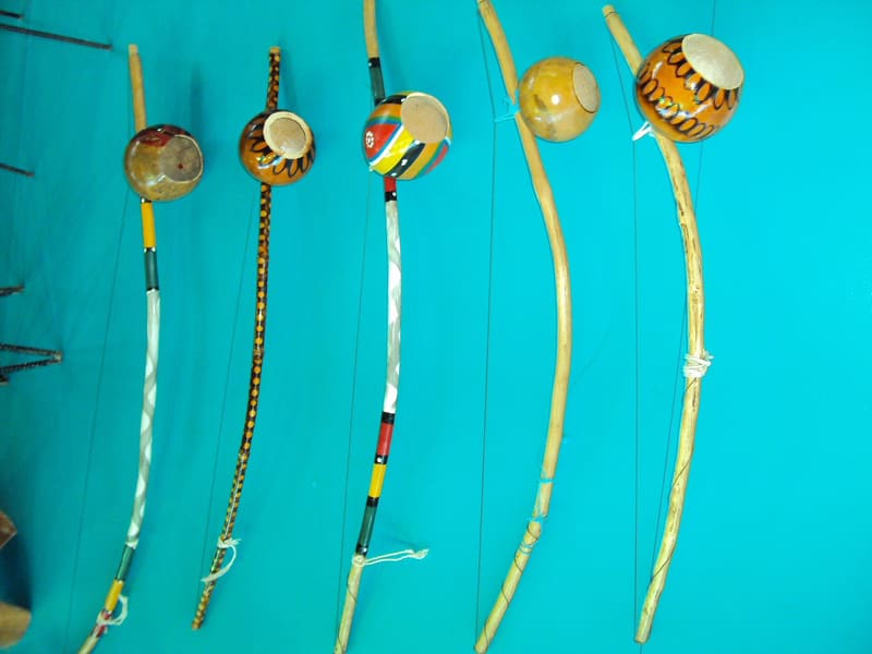 Five berimbaus laid side by side against a turquoise surface, each a curved wooden bow strung with a thin wire and fitted with a gourd resonator, some painted in bright patterns and others left natural.