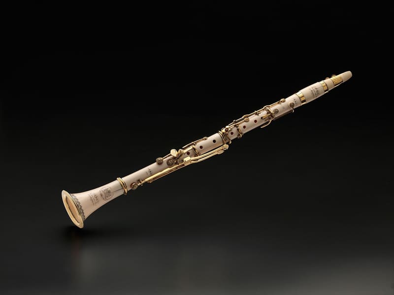 Ivory-coloured clarinet with gold keys, rings and engraved decoration, angled diagonally with the bell at lower left, against a dark background.