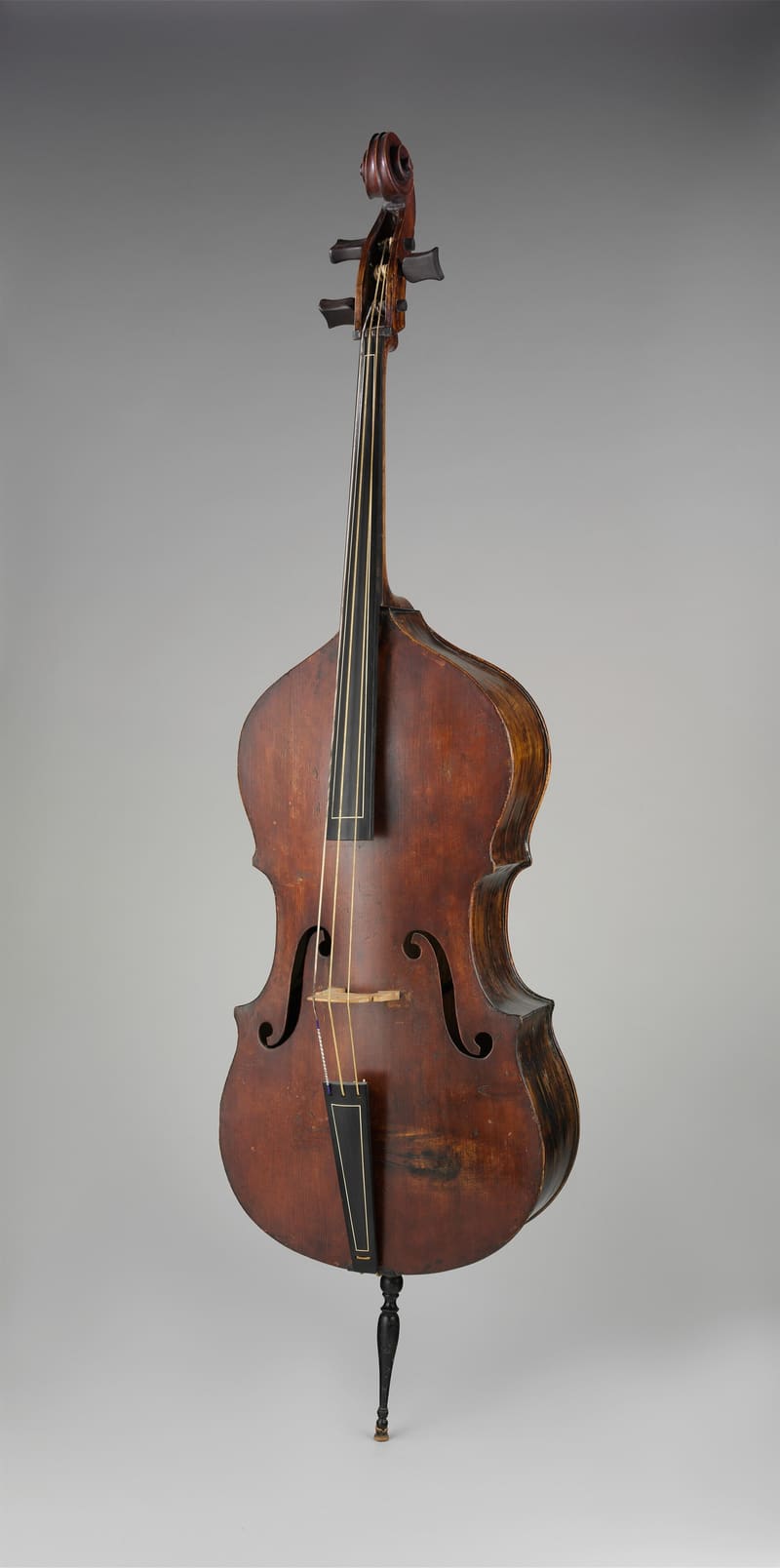 Three-quarter view of an aged reddish-brown double bass with sloping shoulders, f-holes, ebony tailpiece and fingerboard, three strings and a turned black endpin, against a grey background.