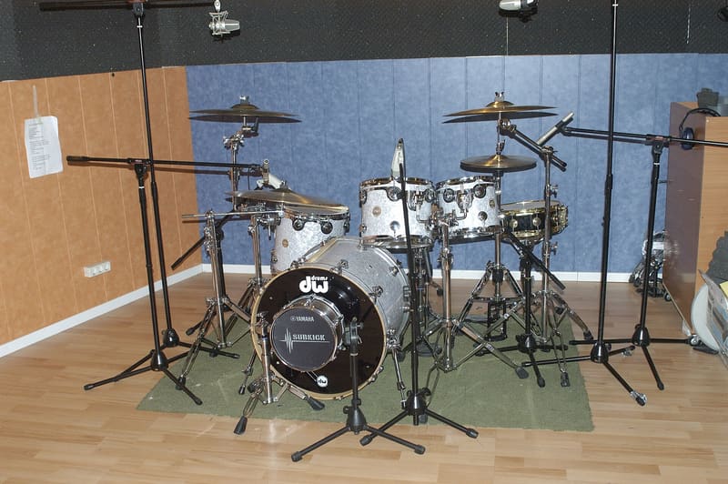 A pearl-white drum kit on a green rug in a recording studio, with bass drum, toms, snare, hi-hat and cymbals surrounded by microphone stands.