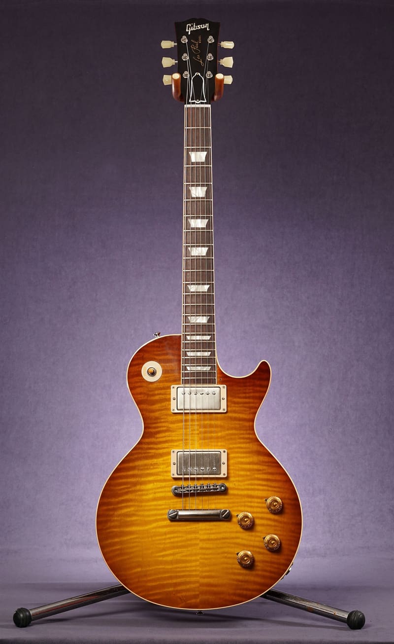 Sunburst solid-body electric guitar with a flame-maple top, two humbucking pickups, four gold control knobs and a long rosewood neck, standing on a metal stand against a purple studio backdrop.