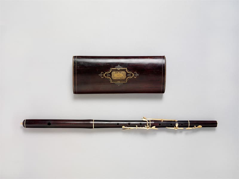 Dark wooden transverse flute with gold rings and gold keys lying horizontally below its matching rosewood case with a gold inlaid cartouche, on a light grey background.