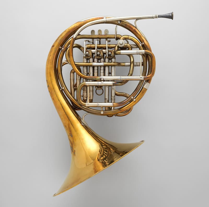 A German double horn in F and B-flat, with coiled brass tubing, three rotary valve levers, nickel-silver slides and a wide bell, against a pale grey background.