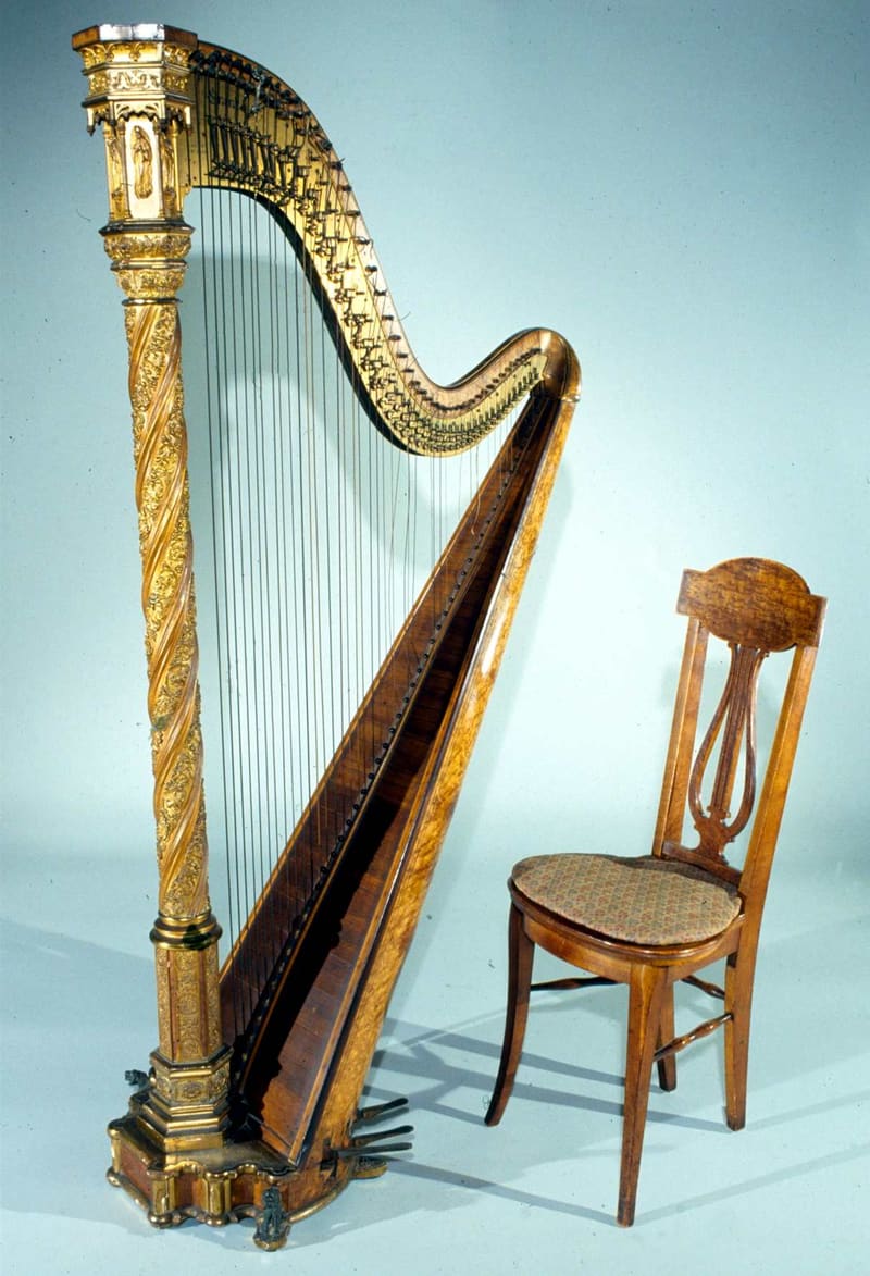 Gothic-style pedal harp with a gilded, carved twisting column, figured-wood soundboard and curved neck with many strings and mechanism discs, displayed beside a small wooden chair on a pale blue background.