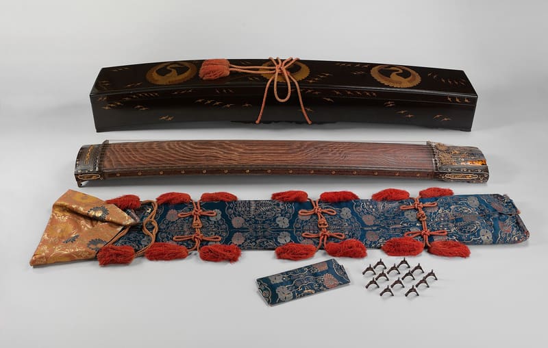 Japanese koto laid out with its black lacquer storage case decorated with gold cranes above it and a brocade cover, spare bridges and a small pouch below, all on a pale grey surface.