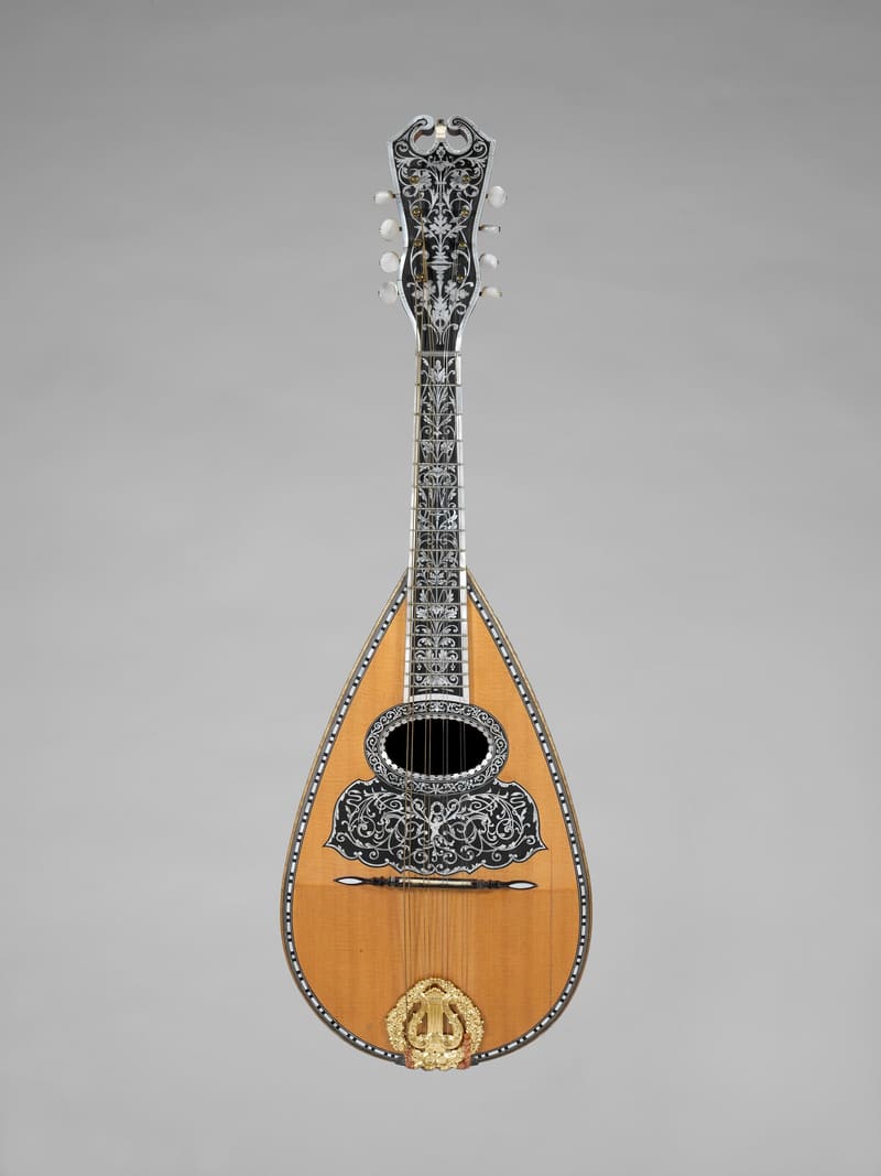 Bowl-back mandolin with a spruce top, ornate black fingerboard and pegbox inlaid with mother-of-pearl, a gilt lyre-shaped tailpiece and eight strings, shown upright from the front on a grey background.