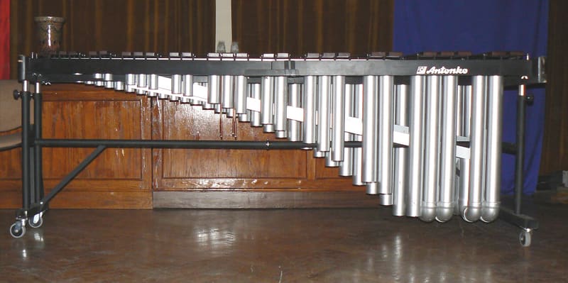 Concert marimba on a wheeled black frame, with rosewood bars along the top and graduated silver resonator tubes hanging beneath, photographed from the side on a stage.