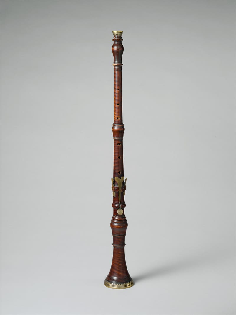 A slender baroque oboe of reddish-brown turned wood with a flared bell, brass-trimmed ends and a single brass key, standing upright against a plain grey background.