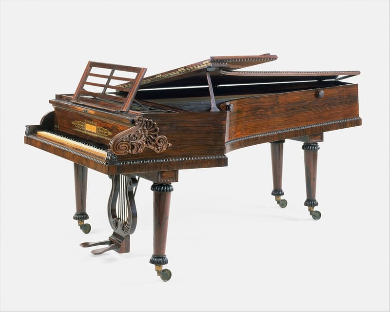 Rosewood grand piano with carved scroll cheek blocks, tapered turned legs on brass casters and a lyre-shaped pedal assembly, shown from the keyboard end with the lid and music desk raised against a plain white background.
