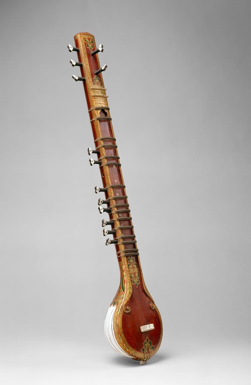 Sitar with a long hollow neck painted red with gold floral borders, movable frets tied with gut, ivory-coloured tuning pegs and a round gourd resonator, shown at an angle against a grey studio background.