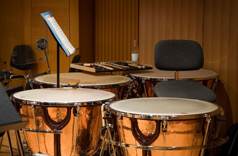 Four copper-bowled timpani with pale calfskin heads, pedals and tuning rods on a wood-panelled stage, with mallets on a tray, a music stand and a chair behind.