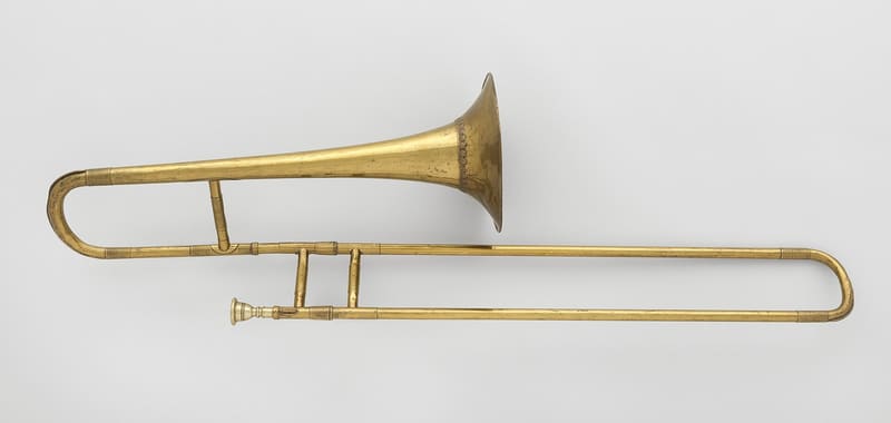 A mid-19th-century brass trombone with a double slide, a small mouthpiece at left and a flaring bell, lying horizontally against a pale grey background.