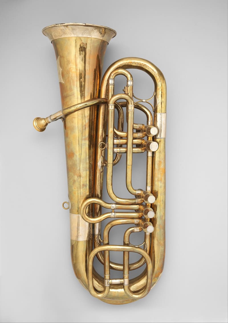 A large mid-19th-century German brass tuba with a wide engraved bell, silver-coloured bands and five valves with looped tubing, shown upright against a soft grey background.