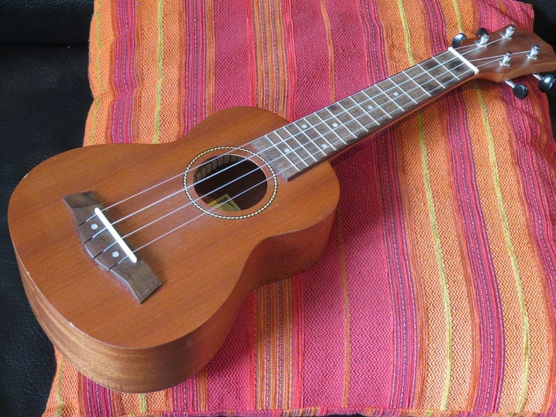 Soprano ukulele in laminated mahogany with four strings, a rosette-edged soundhole and a dark fretboard, lying at an angle on a red and orange striped cushion.