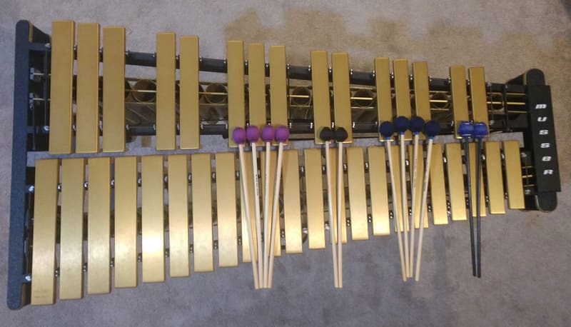 Overhead view of a vibraphone with two rows of golden aluminium bars over metal resonator tubes and several pairs of yarn-wrapped mallets resting across the bars, on a carpeted floor.